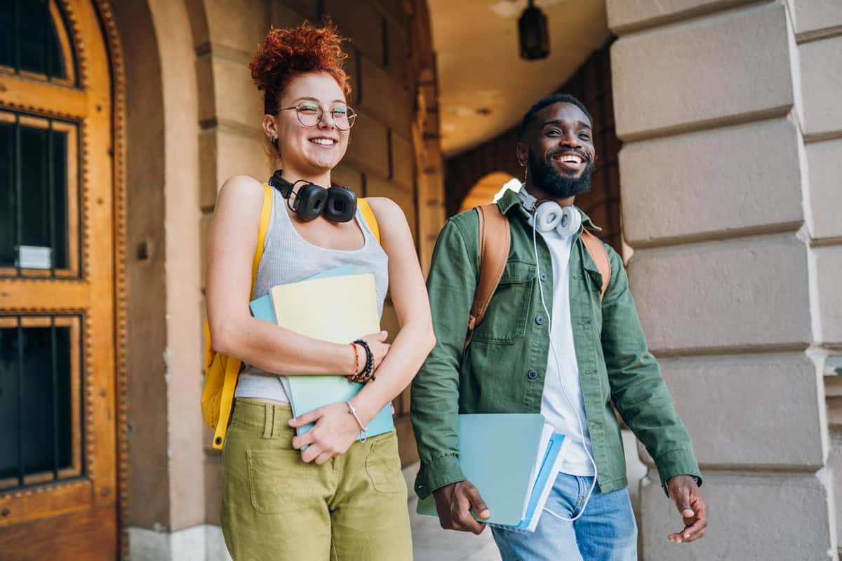 Two smiling students with headphones and notebooks walk together outside a building.