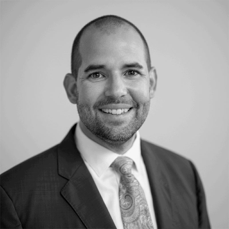 Smiling man in a suit and patterned tie against a plain background. Black and white photo.