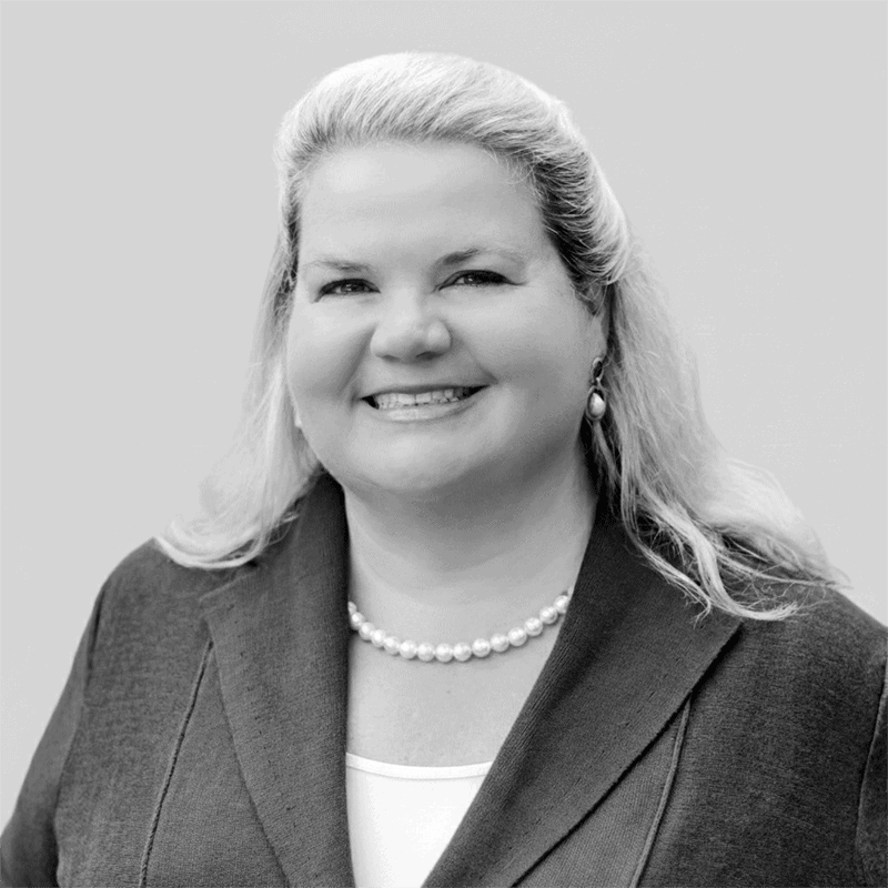 Smiling woman with long blonde hair, wearing a blazer, white top, pearls, and earrings, on a plain background.